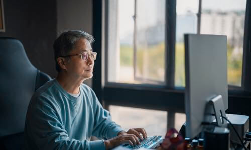 Diverse older man sitting at a desk and looking at his computer monitor while typing