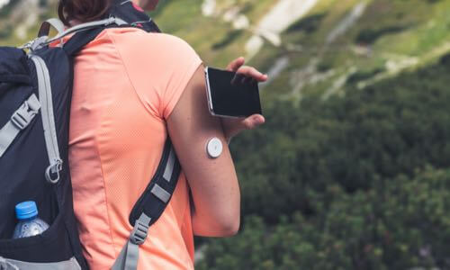 Female hiker outdoors wearing backpack & placing CGM reader by sensor to get glucose reading