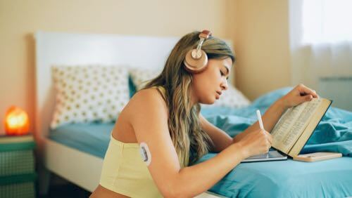 Young woman with CGM on her upper arm leaning over her bed, reading a book & using a tablet
