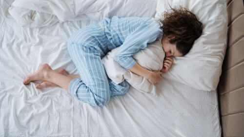 Aerial view of young woman wearing pajamas and curled up & hugging a pillow in bed