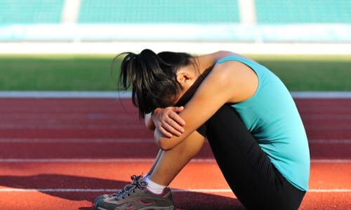 Young female athlete sitting on running track, resting her head over her arms while hugging her knees