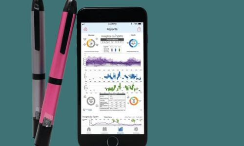 Gray & pink smart pens standing next to a smartphone showing insulin tracking data